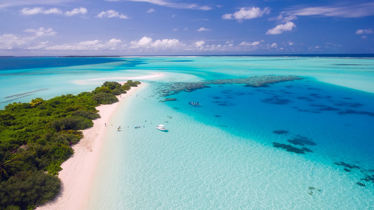 Aerial view of a green island with a white sand beach beside shallow turquoise water, a small boat and a few swimmers.